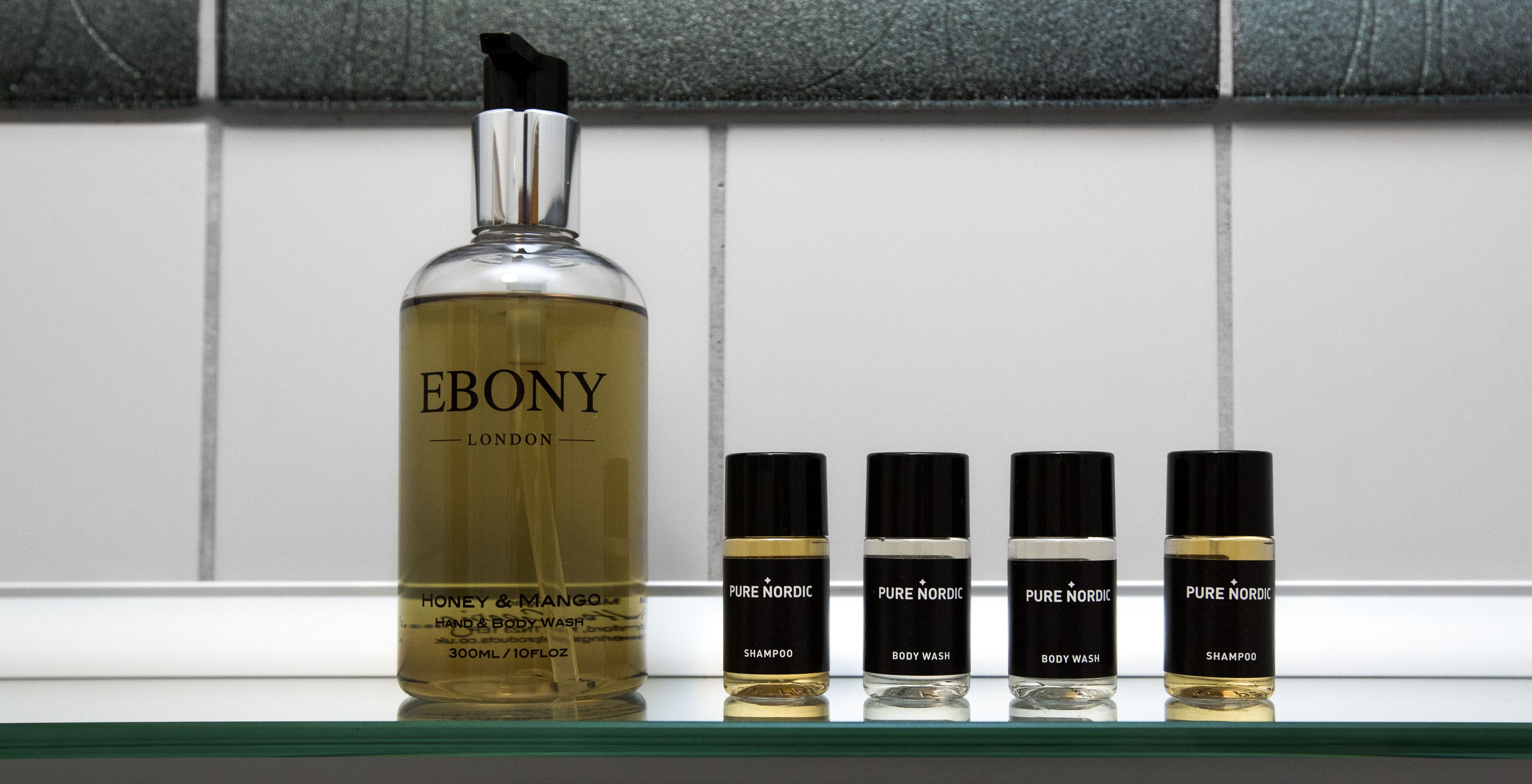 Una bottiglia di detergente corpo e mani Ebony London Honey & Mango e quattro flaconi di detergente corpo Pure Nordic su uno scaffale.