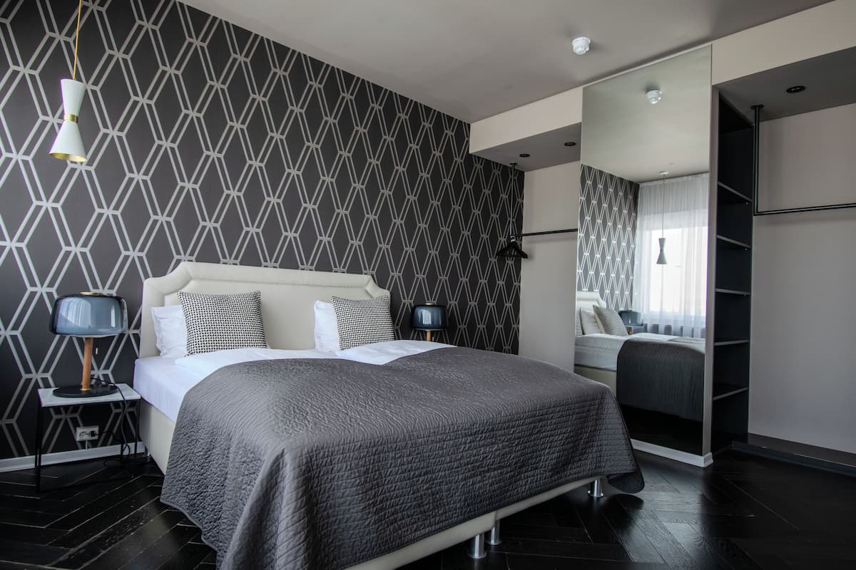 Quarto moderno com parede estampada em padrão geométrico, cama grande com colcha cinza, mesas de cabeceira com luminárias e guarda-roupa com espelho.