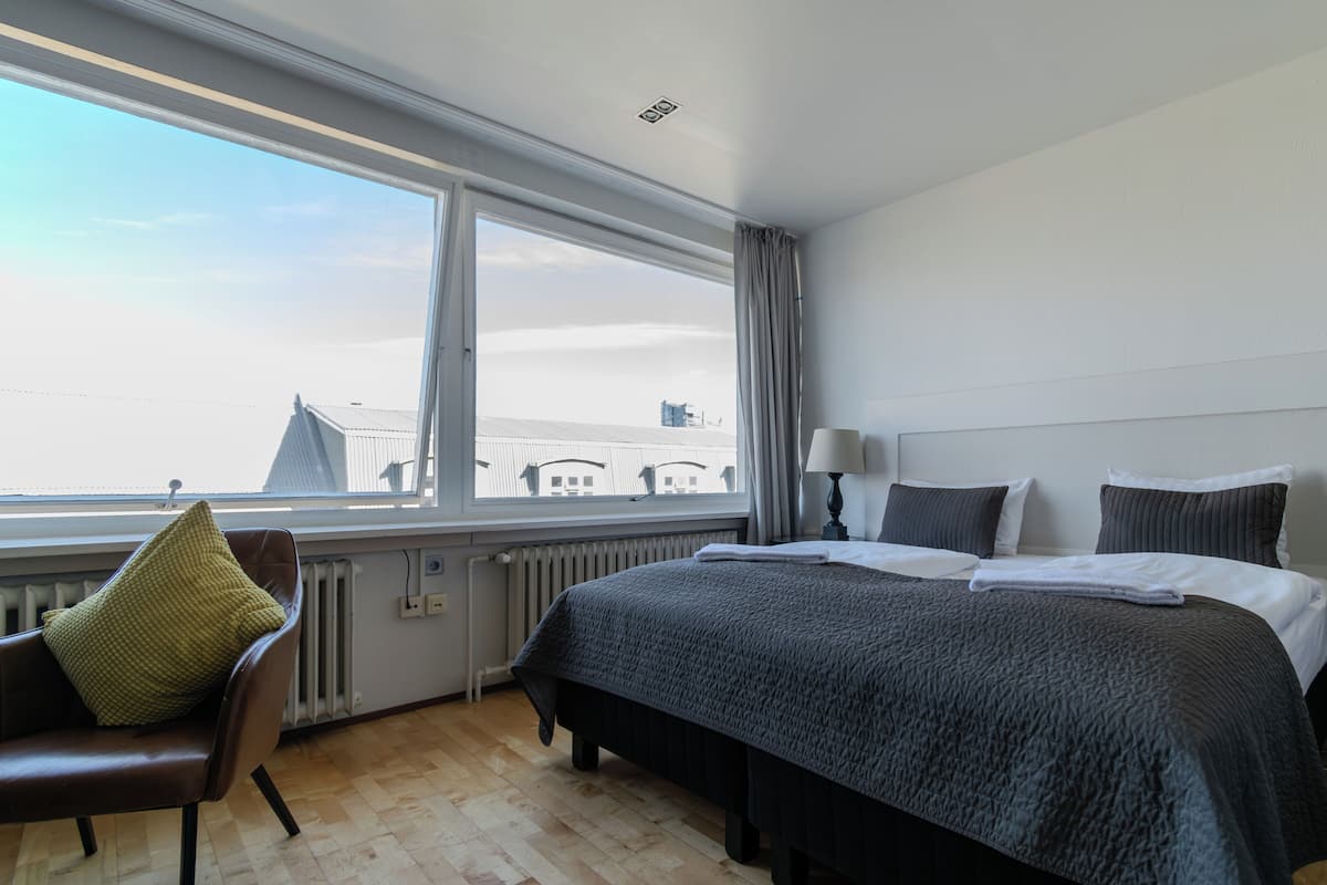 Quarto de hotel com uma cama grande, uma cadeira, uma mesa de cabeceira e uma janela com vista para edifícios.