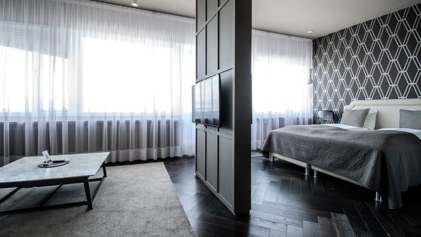 A modern hotel room with a large bed, a flat-screen TV mounted on a wall, a small table, and a view of the city through sheer curtains. - Máni Apartments by Heimaleiga (Reykjavik)