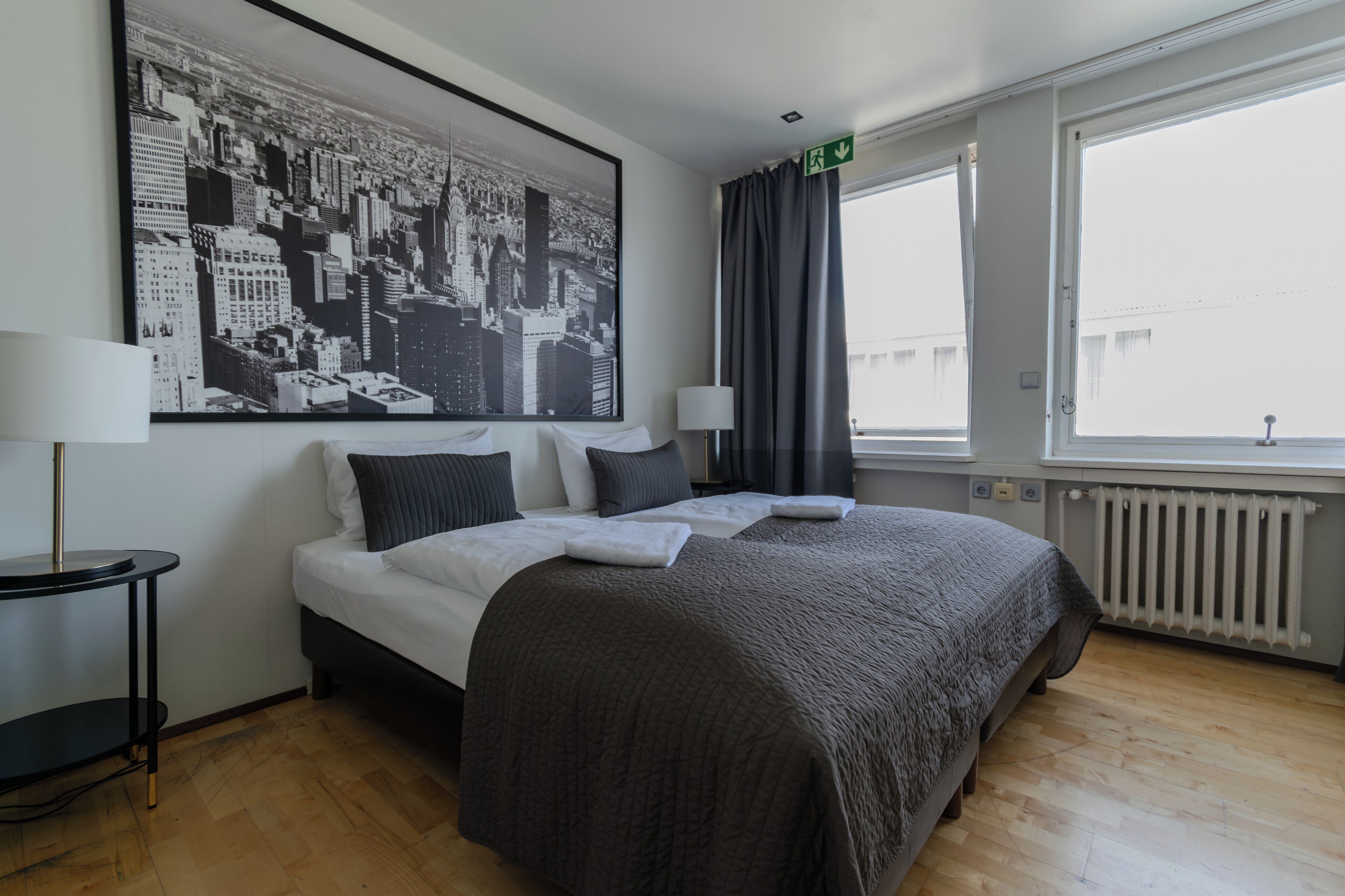 Una habitación de hotel con una cama grande, una mesita auxiliar, una lámpara, una ventana con cortinas y un cuadro de un paisaje urbano en la pared.
