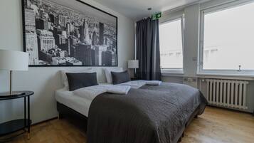Una habitación de hotel con una cama grande, una mesita auxiliar, una lámpara, una ventana con cortinas y un cuadro de un paisaje urbano en la pared.