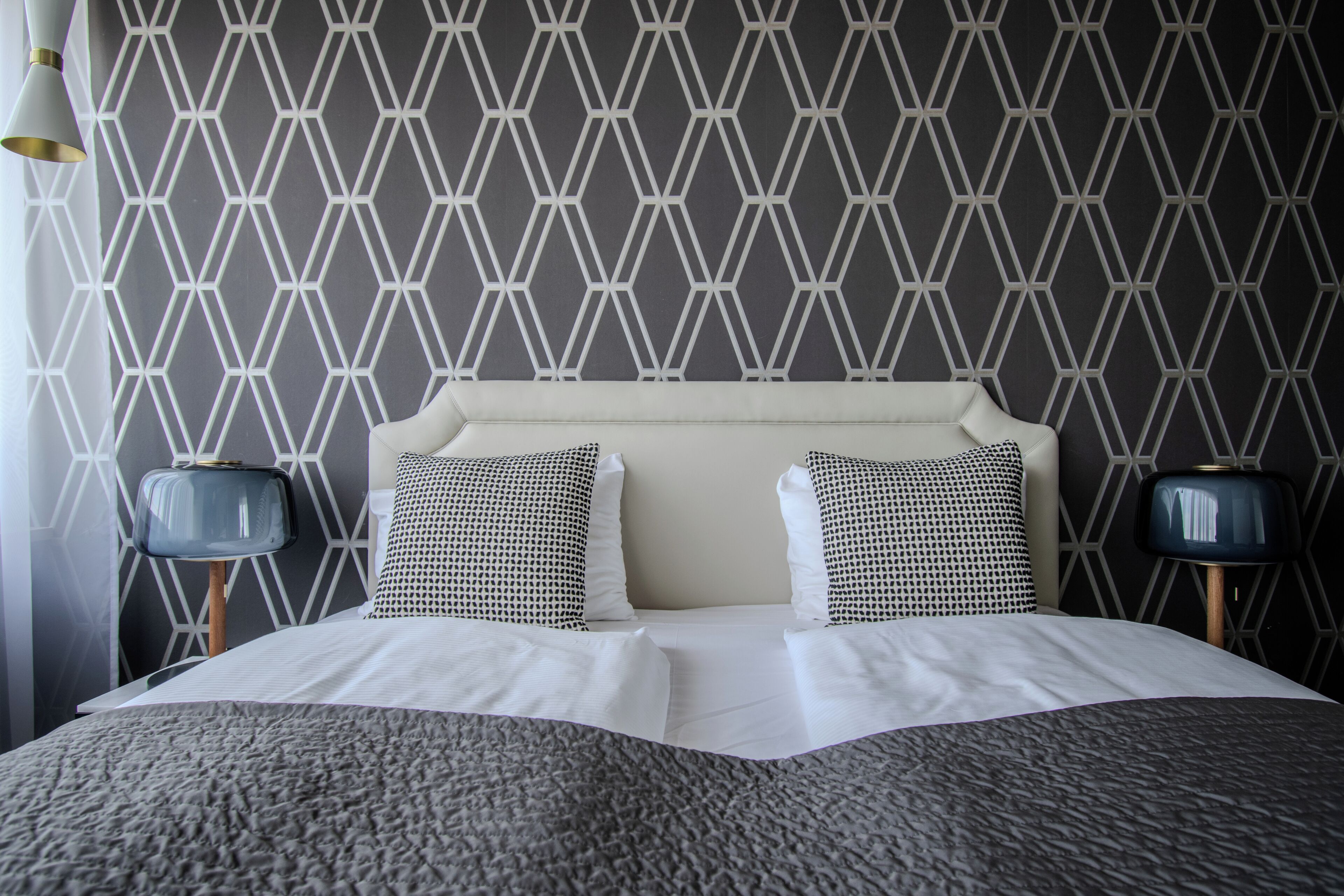 Cama bem arrumada com cabeceira estampada em padrão geométrico, duas luminárias de cabeceira e um cobertor texturizado.