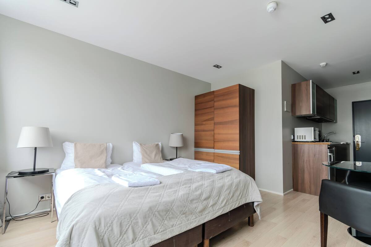 Quarto de hotel moderno com cama bem arrumada, guarda-roupa de madeira, kitchenette e mesa de vidro.