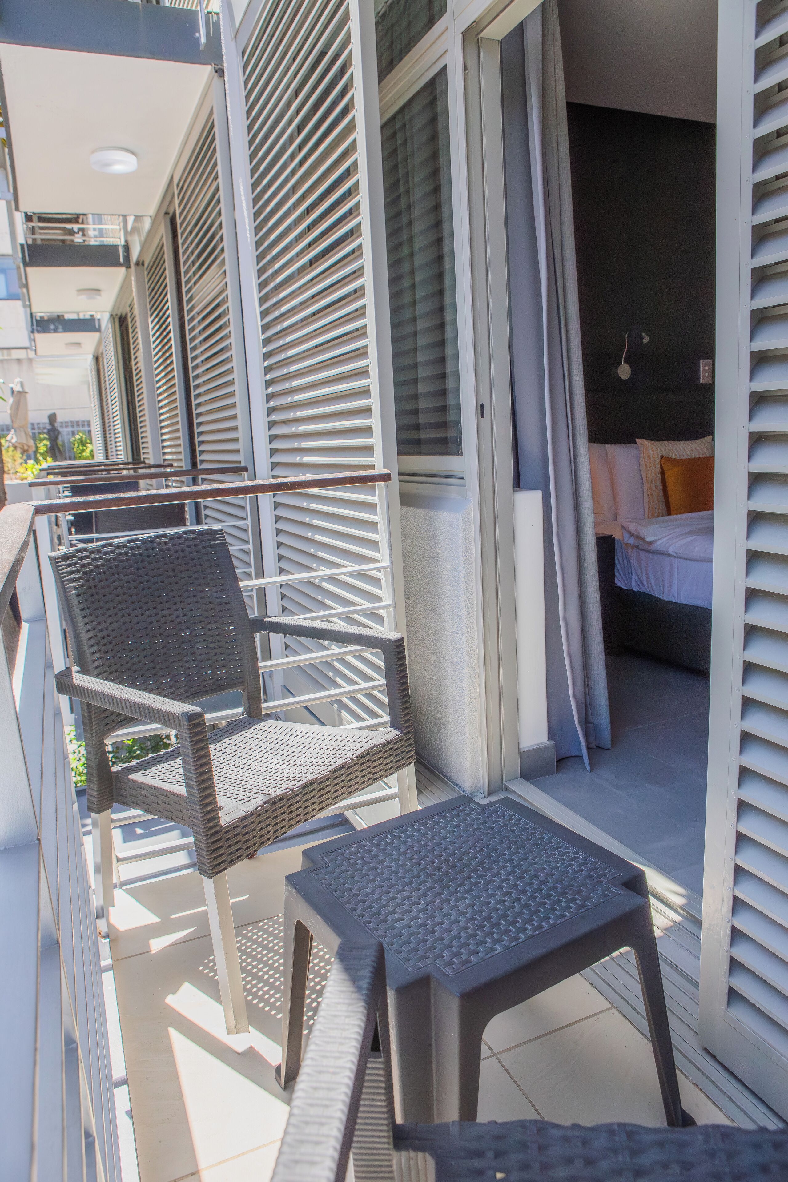 A balcony with wicker chairs and a small table, a bed with white bedding, and a dark-colored wall.