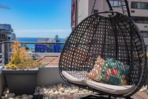 Ein schwarzer Korbsessel mit Kissen auf einer Dachterrasse mit Blick auf das Meer und Gebäude.