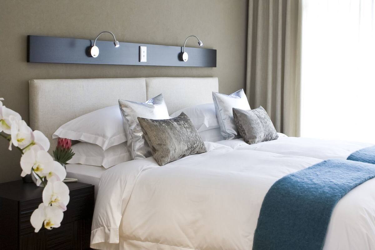 Un lit bien fait, avec du linge de lit blanc et gris, un plaid bleu et un luminaire mural moderne fixé au-dessus.