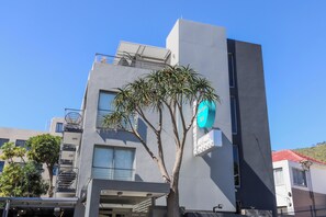 Ein modernes Gebäude mit einer grauen Fassade, einem Balkon mit Metallgeländer und einem Baum davor.