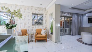 Een moderne hotel lobby met een glazen salontafel, twee fauteuils, een potplant en een grote spiegel.