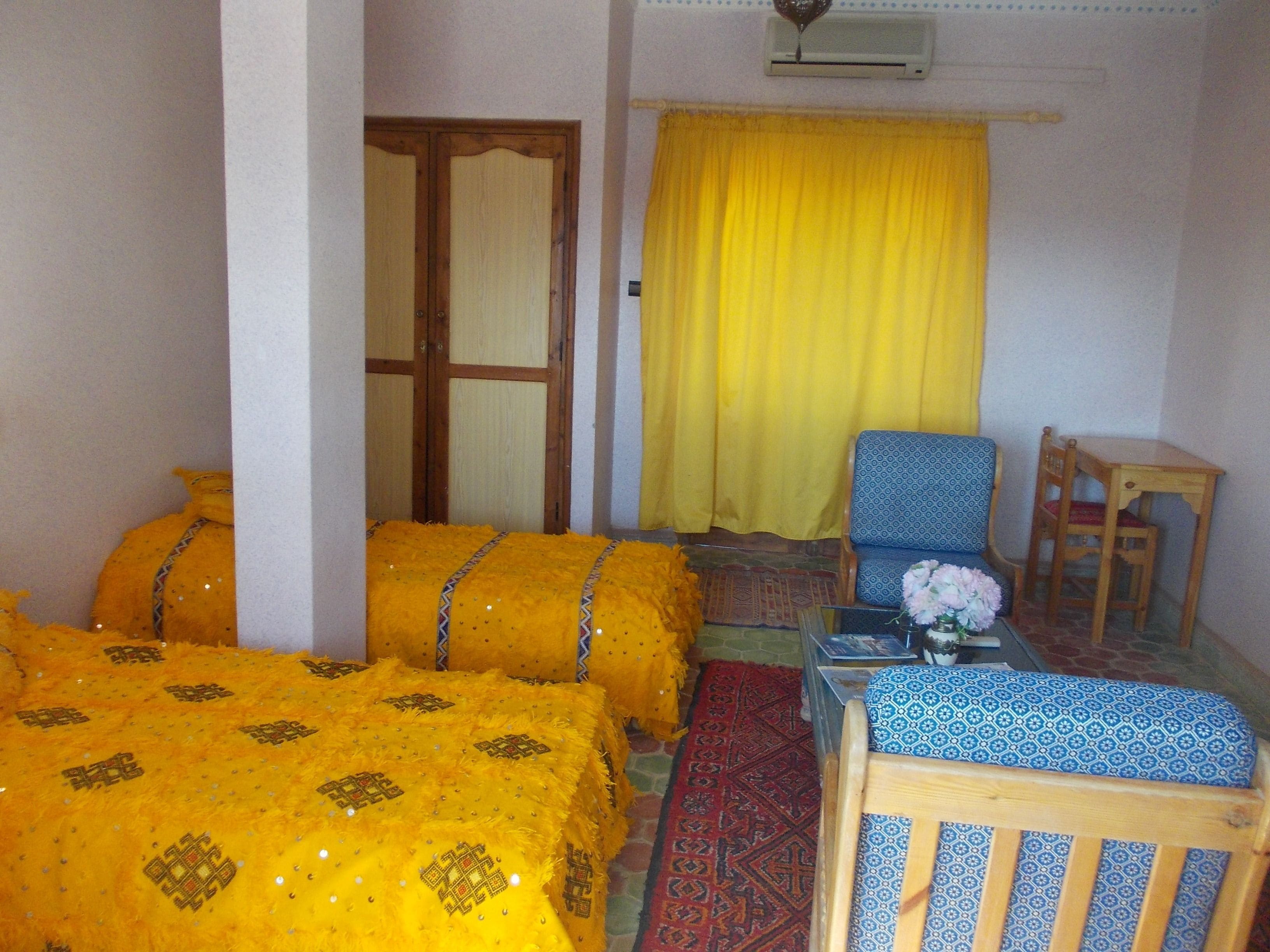 A room with two beds covered in yellow bedding, a blue patterned sofa, a wooden chair, a small table with a vase, and a yellow curtain.