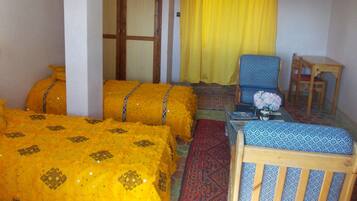 A room with two beds covered in yellow bedding, a blue patterned sofa, a wooden chair, a small table with a vase, and a yellow curtain.