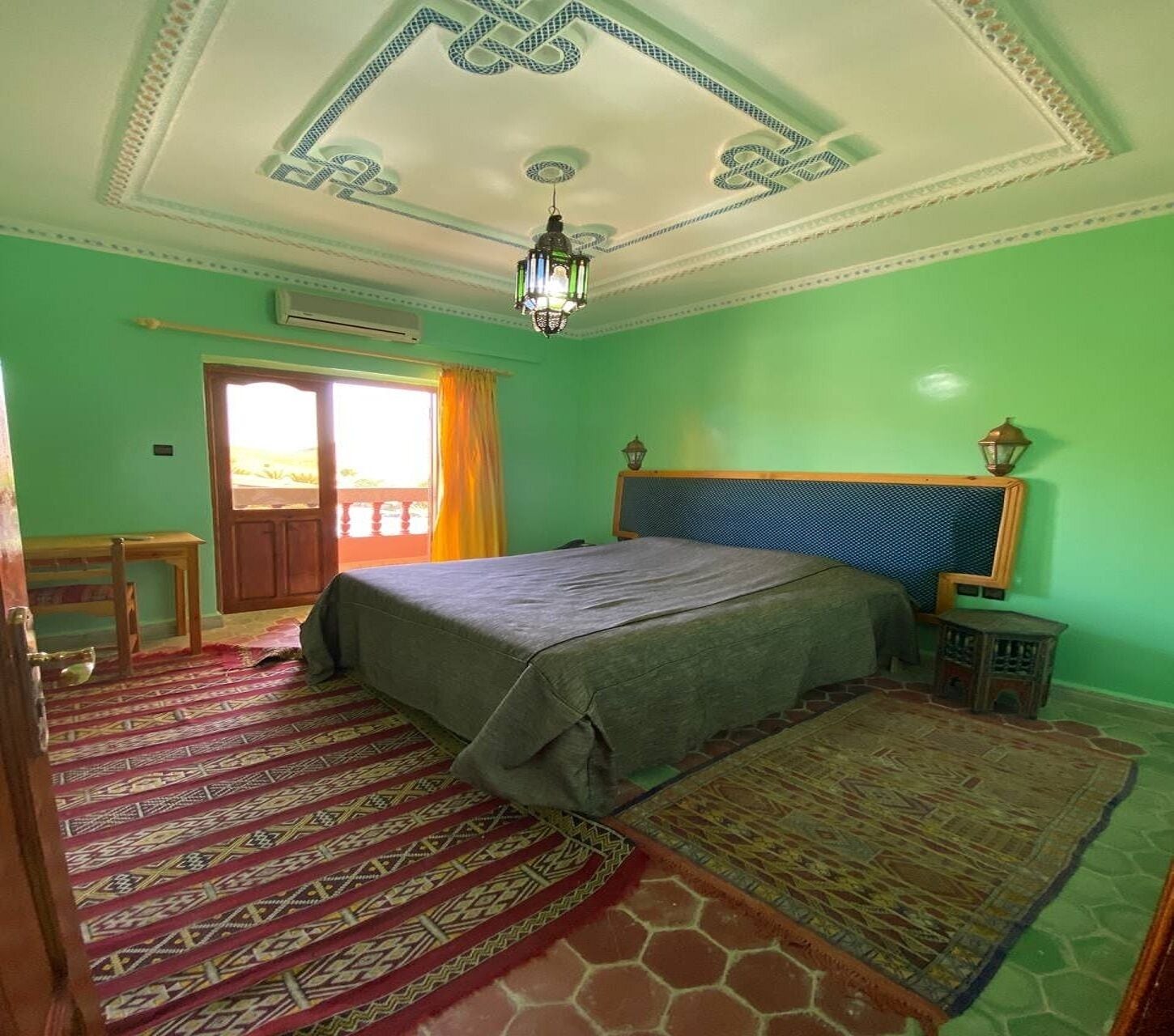 A bedroom with a bed, a patterned rug, a wooden bench, a small table, and a door leading to an outdoor area.