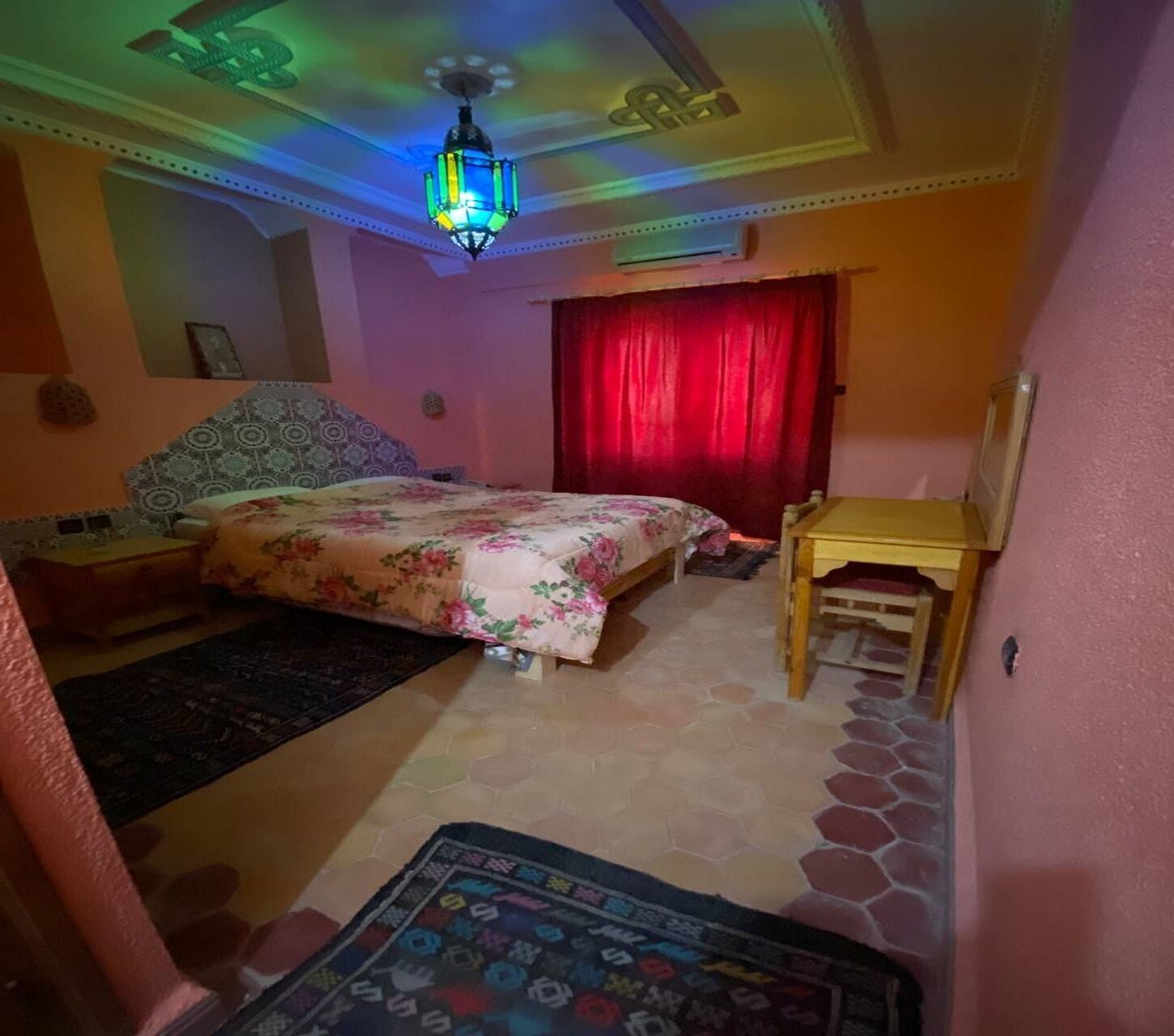 Een slaapkamer met een bed, nachtkastje, houten tafel, rood gordijn en een kamerbreed tapijt met een patroon.