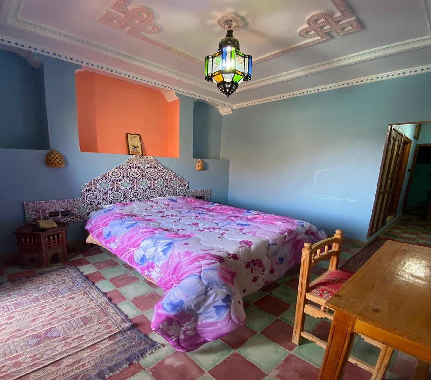 A bedroom with a bed, a wooden chair, a small table, a patterned rug, and a decorative ceiling light.