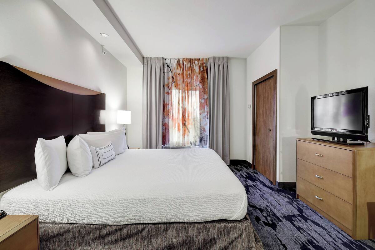 A hotel room with a large bed, a flat-screen TV, a wooden dresser, and a window with curtains.