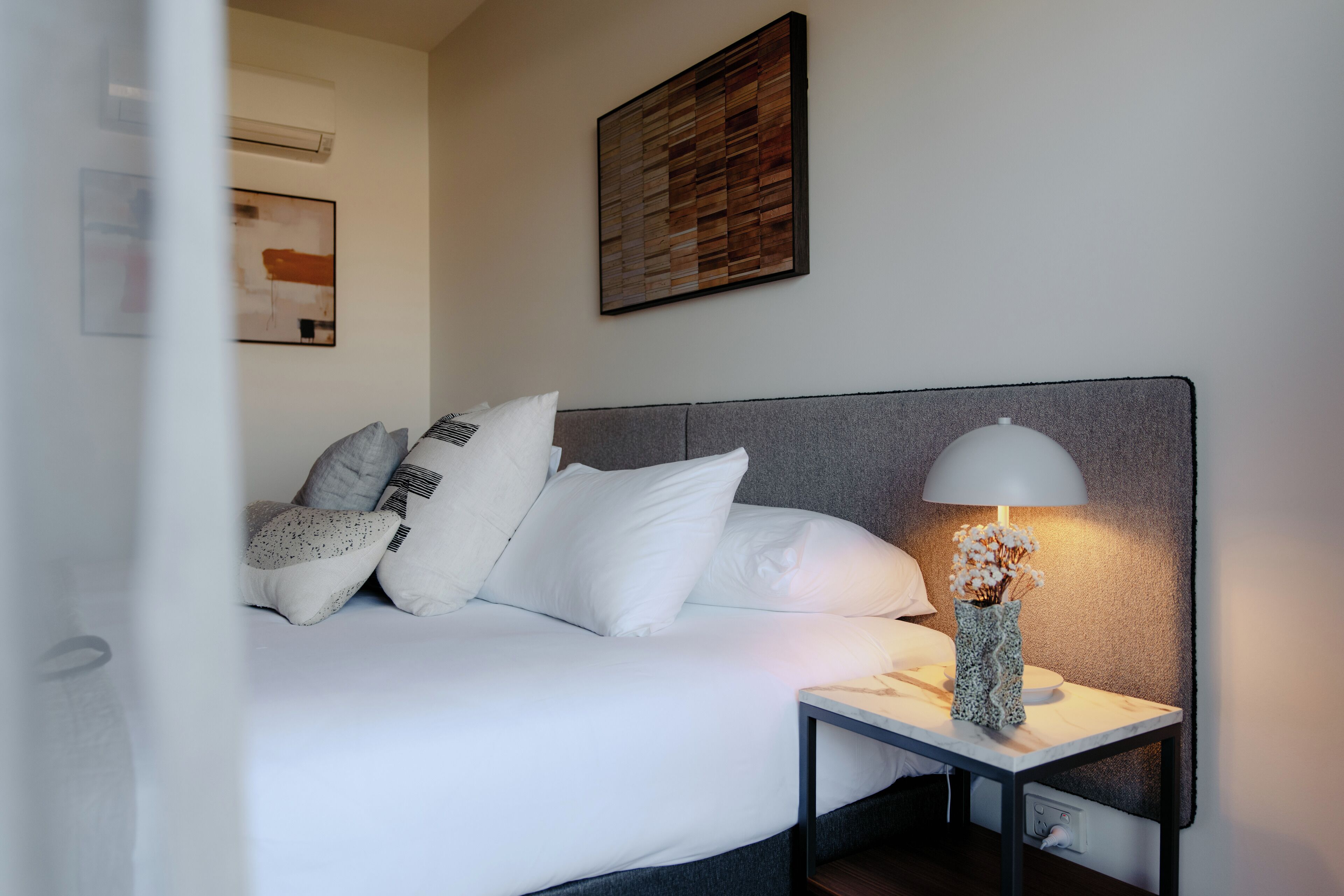 Een keurig opgemaakt bed met witte beddengoed en meerdere kussens, een nachtkastje met een lamp, en twee aan de muur bevestigde kunstwerken.