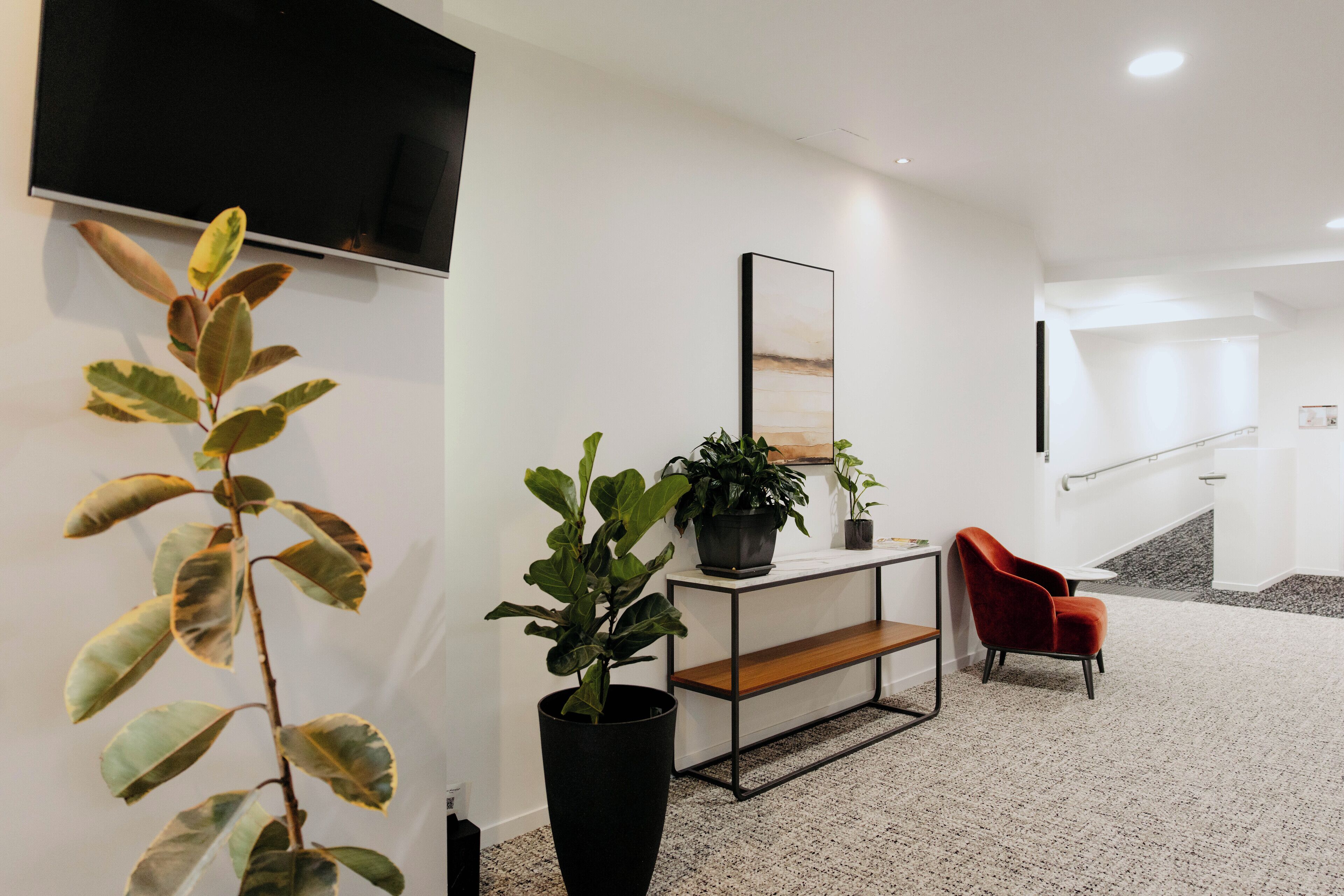 A modern living room with a flat-screen TV, a console table with a potted plant, a red chair, and a staircase in the background.