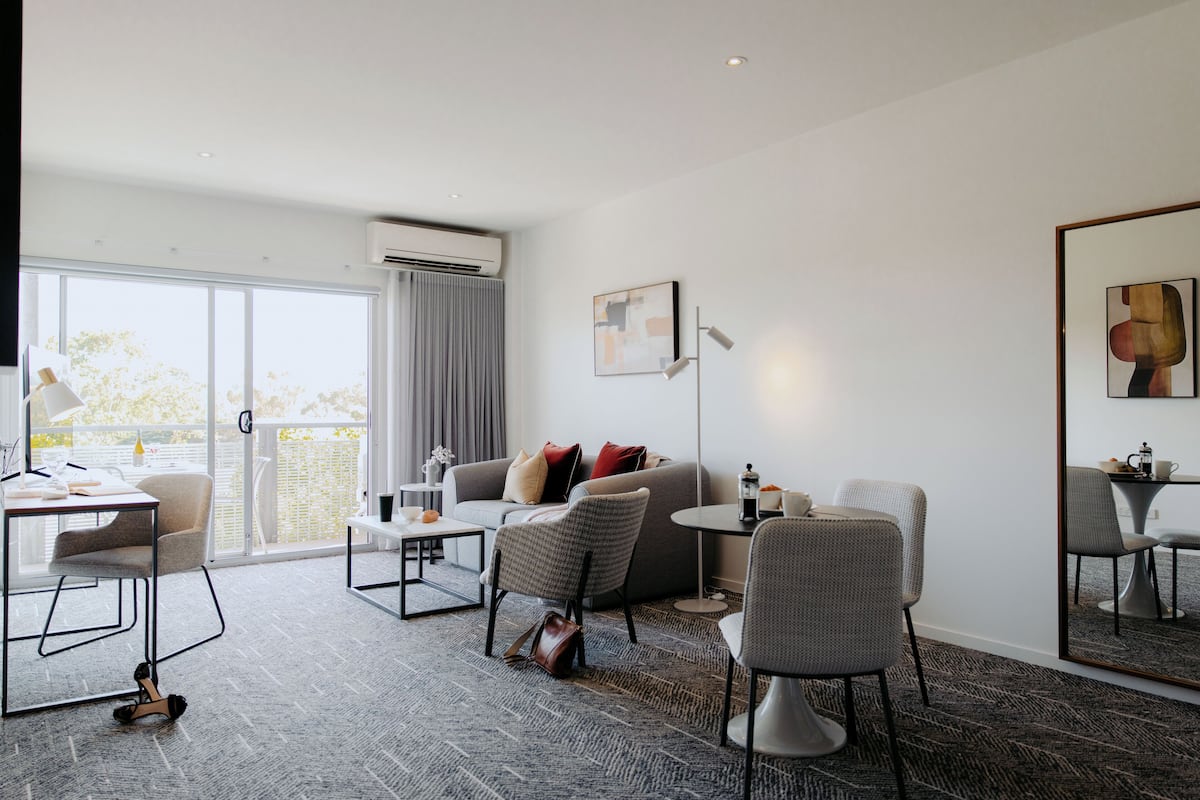 A modern hotel room with a sofa, armchairs, a dining table, and a large window.