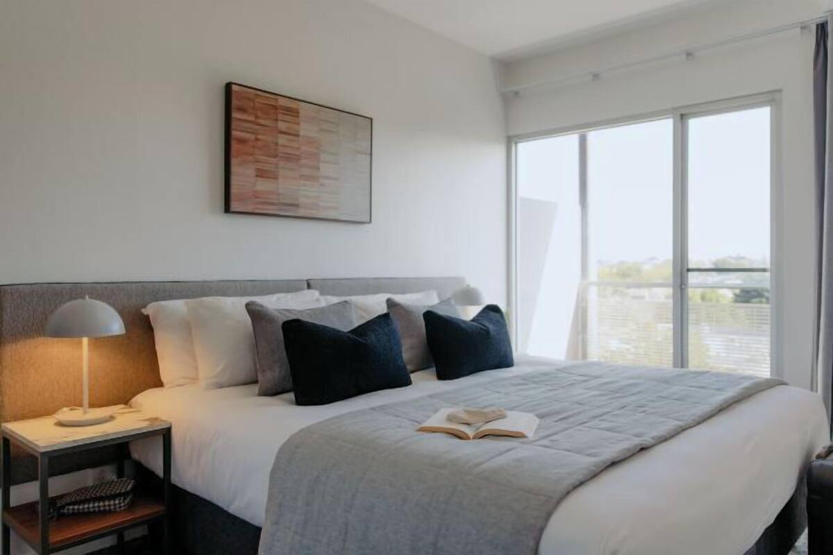 A modern hotel room with a large bed, a nightstand with a lamp, a view of the city through large windows, and a wall-mounted artwork.