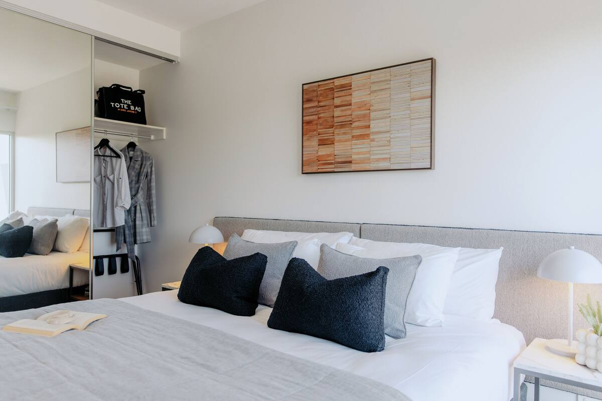 A hotel room with a bed, pillows, a bedside table with a lamp, a wardrobe, and a wall-mounted artwork.