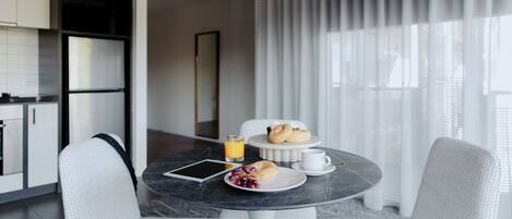 A modern dining area with a marble table, white chairs, a tablet, a plate of grapes, a glass of orange juice, and a small white table with pastries.