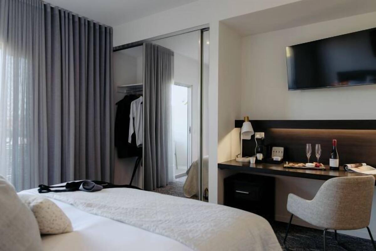 A hotel room with a bed, a desk with a flat-screen TV, a chair, a coat rack, and a sliding door.