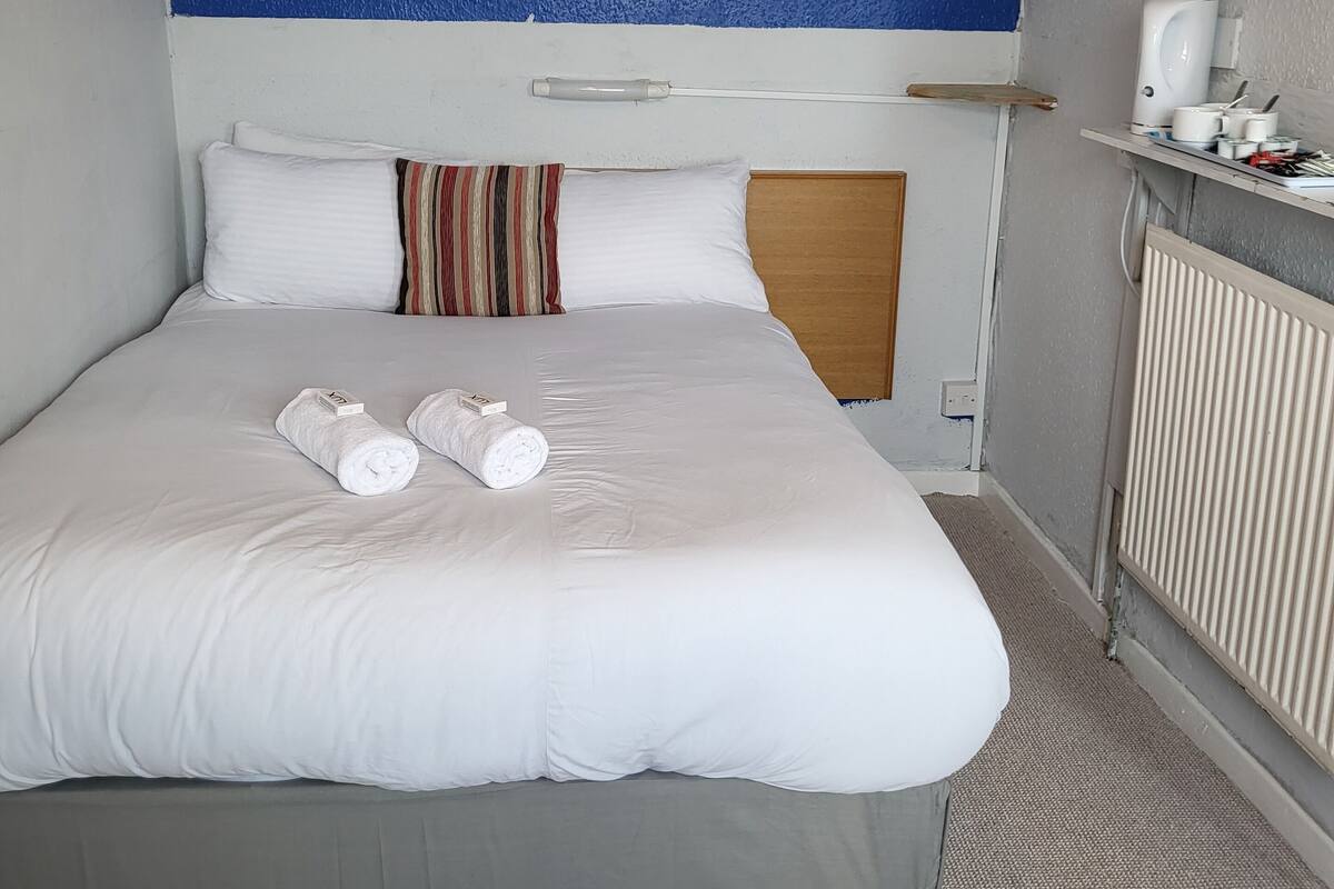 Habitación de hotel con una cama grande, ropa de cama blanca, una pared azul como detalle y una mesita de noche con un hervidor, además de un radiador.