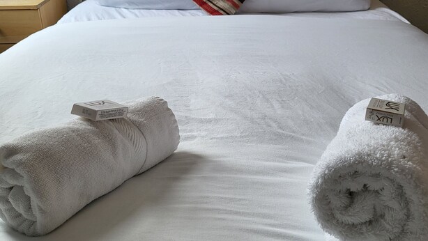 A neatly made bed with white linens, a striped pillow, and two rolled towels with tags.