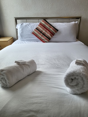 Una cama bien hecha con sábanas blancas, una almohada a rayas y dos toallas enrolladas con etiquetas.
