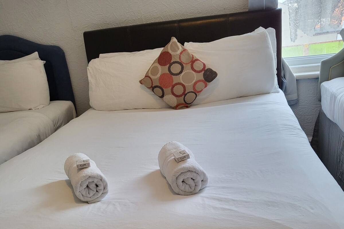 Una cama bien tendida con dos toallas enrolladas y una almohada estampada, un cabecero oscuro y una ventana con vistas a la vegetación.