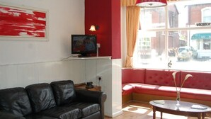 A living room with a black leather sofa, a red accent wall, a television on a stand, a window with a view of the street, and a small table with a vase.