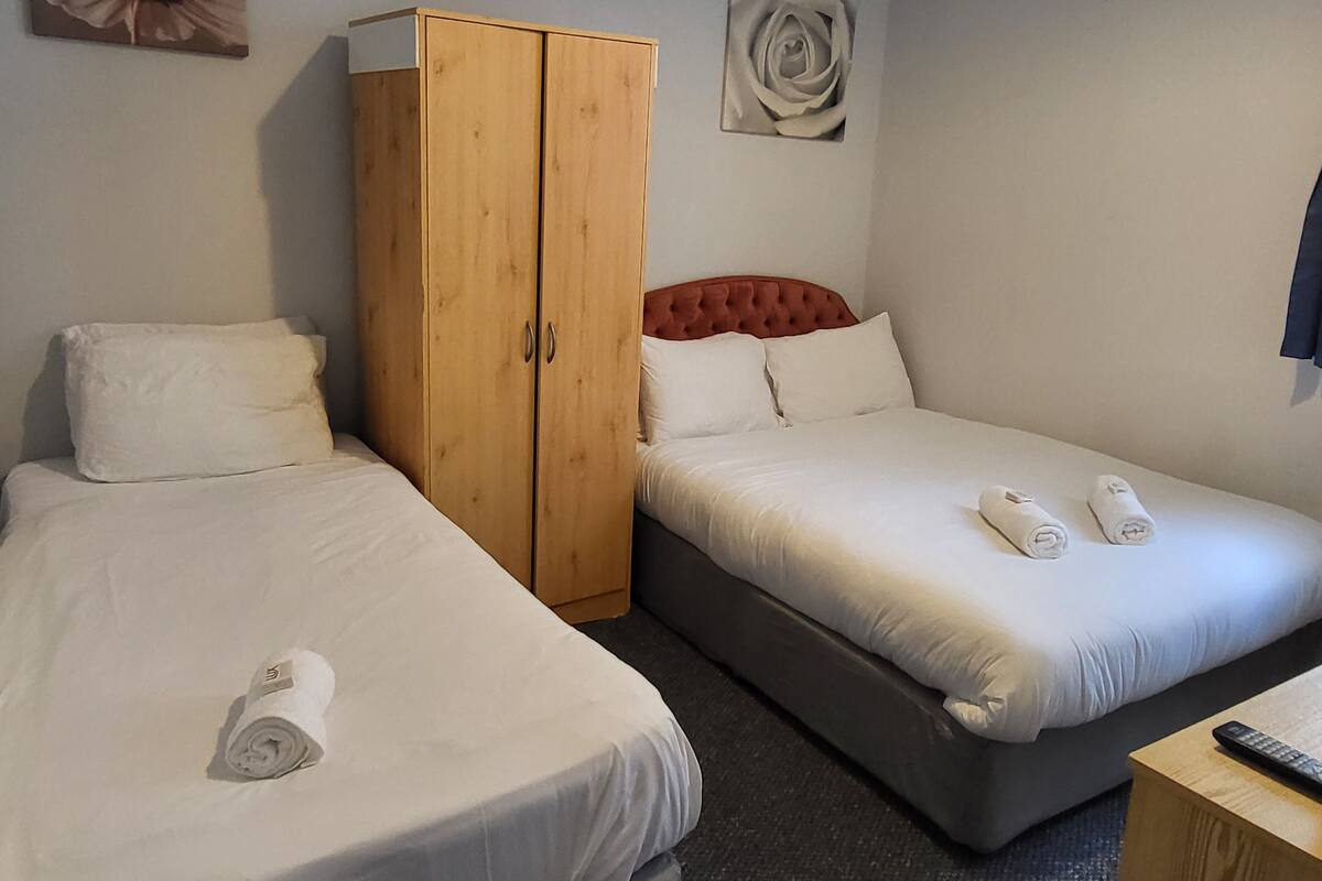 Habitación de hotel con dos camas individuales, un armario de madera, un cabecero rojo y un mando a distancia sobre una mesita de noche.