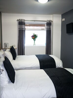 A hotel room with two beds, a window with curtains, a flat-screen TV, and a vase with flowers. - The Maple Hotel (Liverpool)