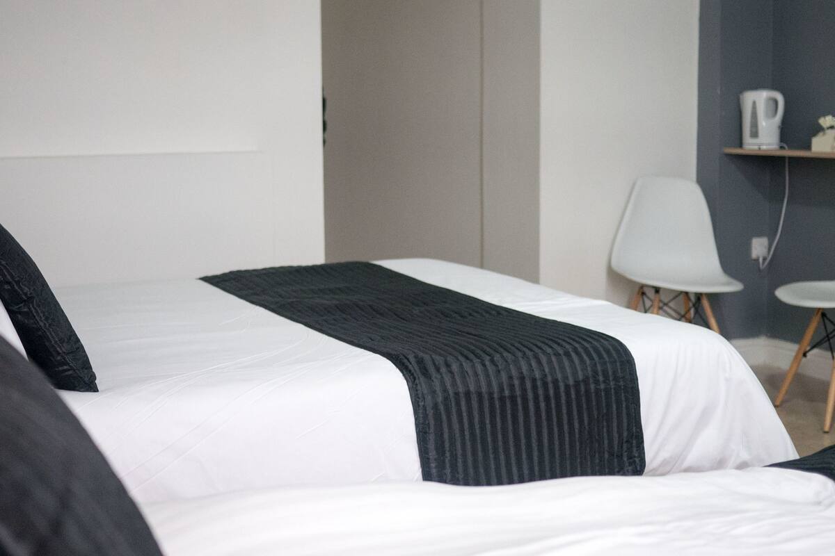 Camera d'albergo con un letto grande, due sedie bianche, un tavolino e uno scaffale con bollitore e tazze.