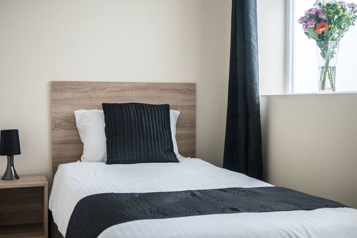Un letto rifatto con lenzuola bianche e nere, una testiera in legno, un comodino con una lampada e una finestra con un'asse di fiori.