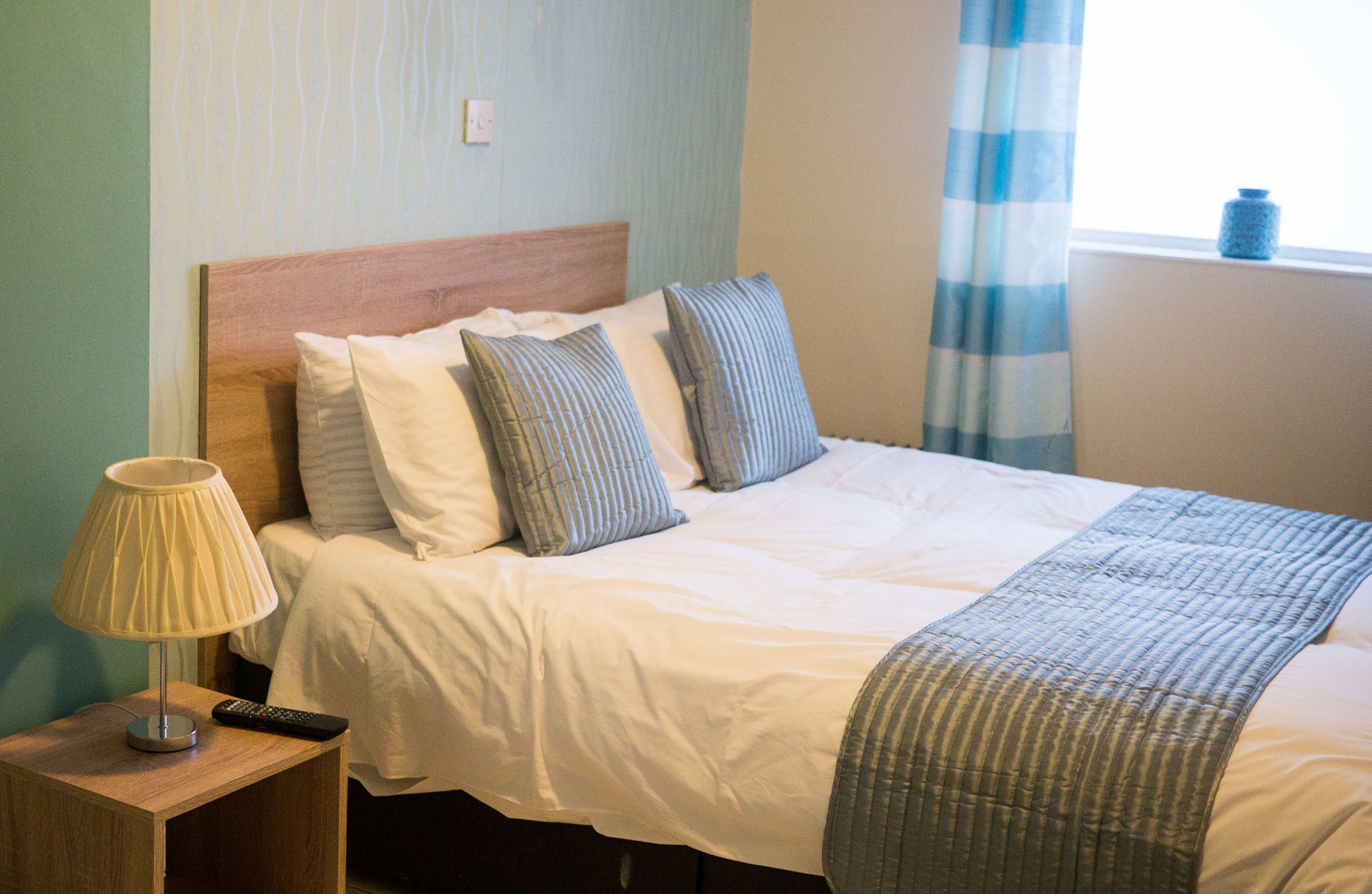 A neatly made bed with a wooden headboard, a bedside lamp, and a remote control on a wooden nightstand.