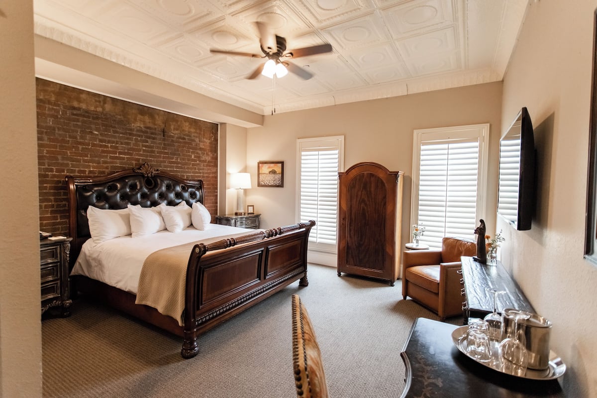 A bedroom with a large bed, a wooden wardrobe, a desk with a tray of glasses, and a ceiling fan.