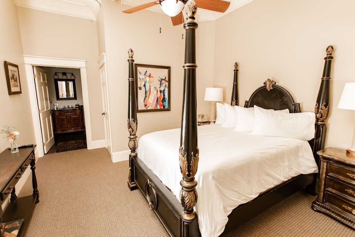A four-poster bed with a dark finish and ornate detailing, a bedside table with a lamp, and a framed painting on the wall.