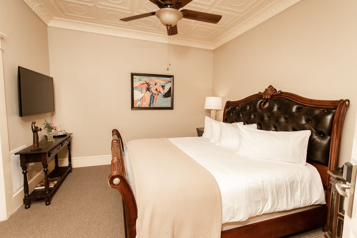 A bedroom with a large bed, a ceiling fan, a painting on the wall, and a nightstand with a lamp.