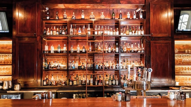 A bar with wooden shelves stocked with various bottles and glasses, a bar counter, and a television screen. - The Grand Hotel (McKinney)
