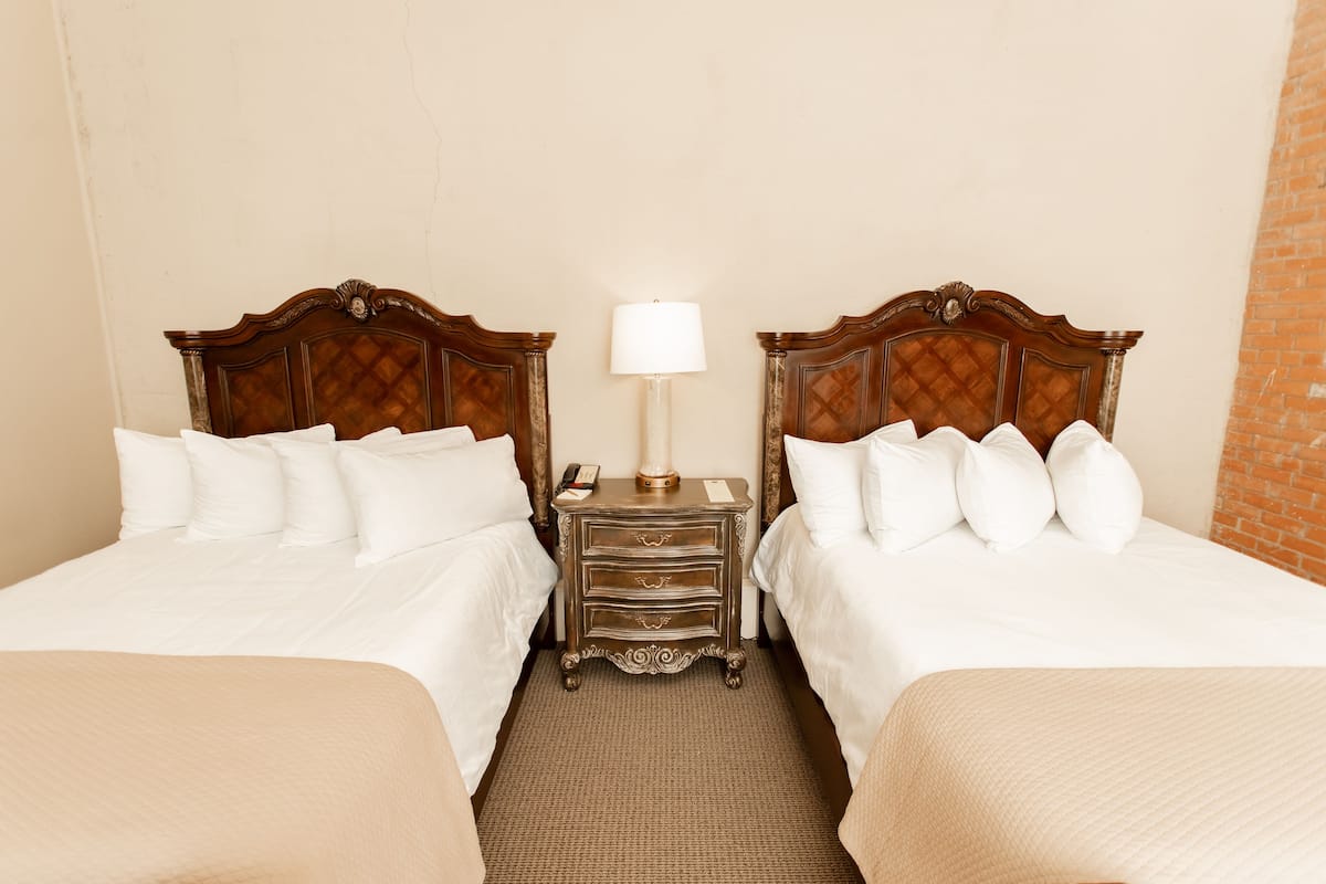 A hotel room with two beds, a nightstand with a lamp, and a brick wall.