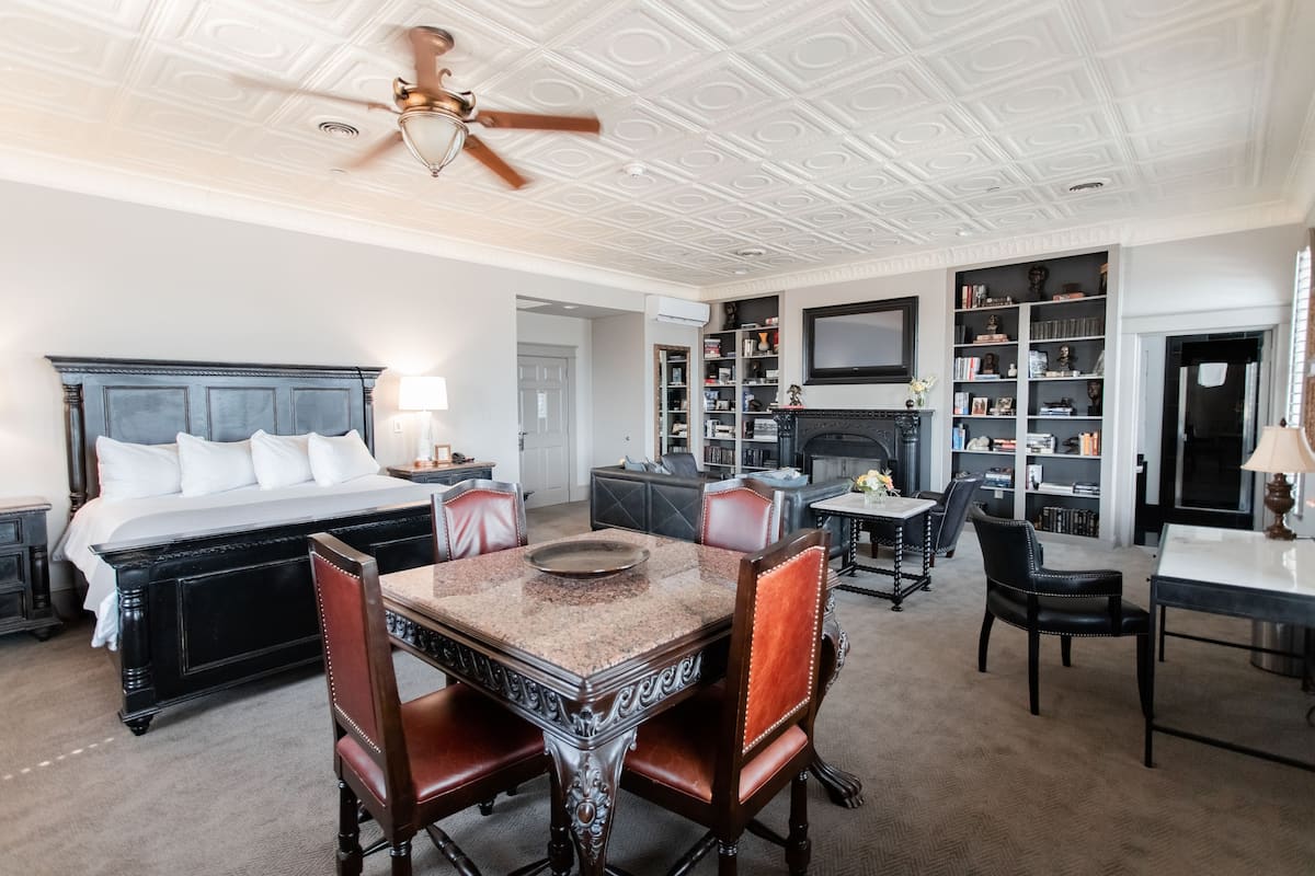 A spacious bedroom with a large bed, a dining area with a table and chairs, a fireplace, and a bookshelf.