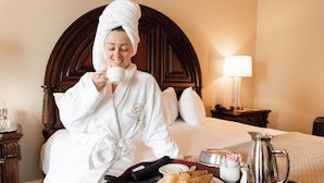 A hotel room with a wooden headboard, a bedside lamp, a tray with breakfast items, and a coffee pot. - The Grand Hotel (McKinney)