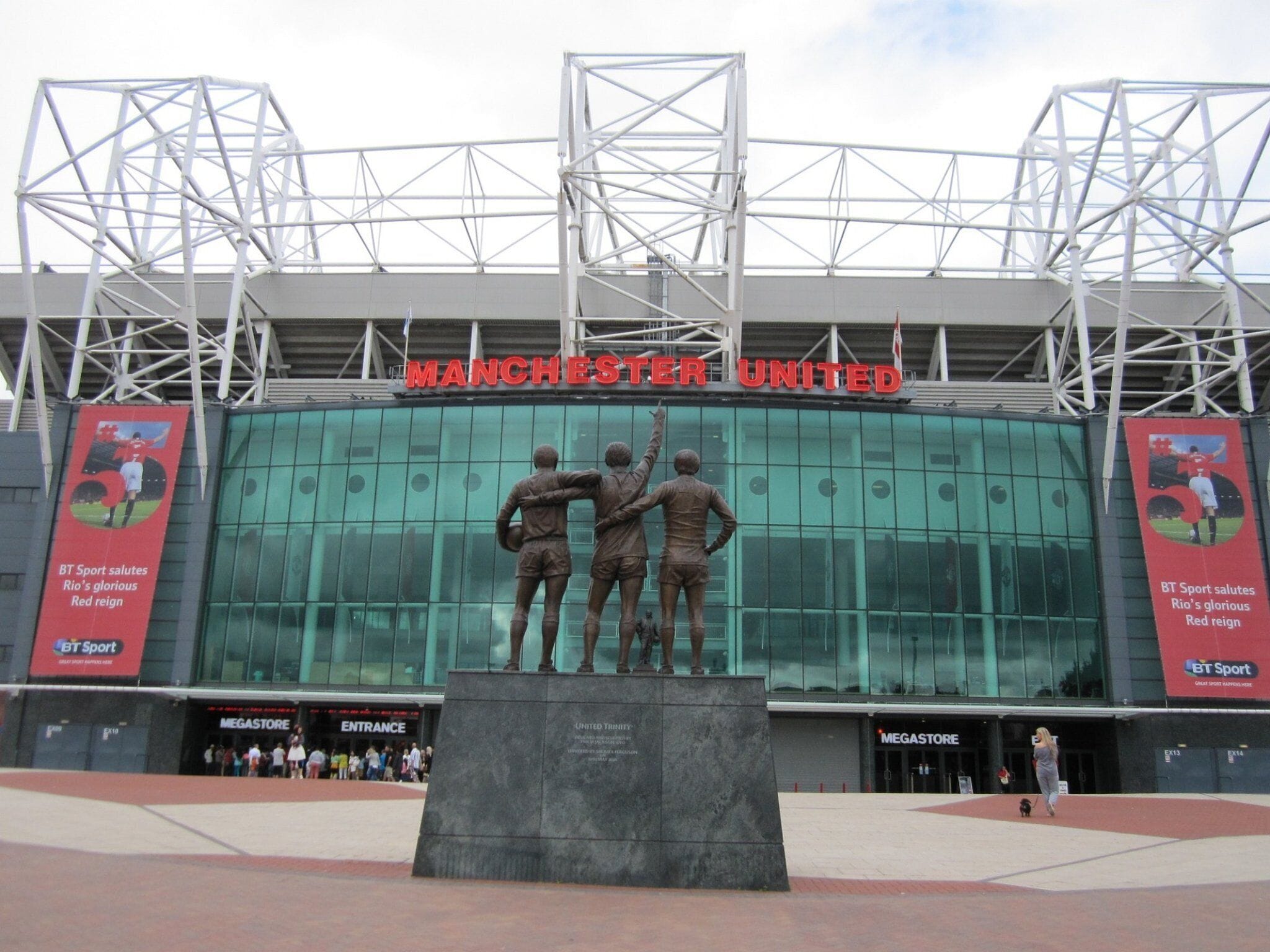 Une entrée de stade avec une grande enseigne sur laquelle on peut lire « MANCHESTER UNITED » et une statue représentant trois personnages.