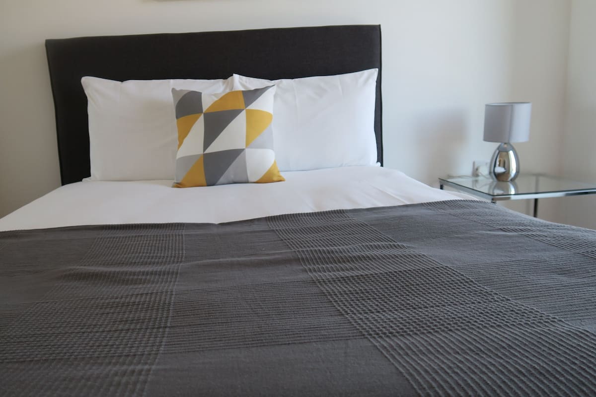 Uma cama bem arrumada com cabeceira preta, roupa de cama branca e um travesseiro com padrão cinza e amarelo.