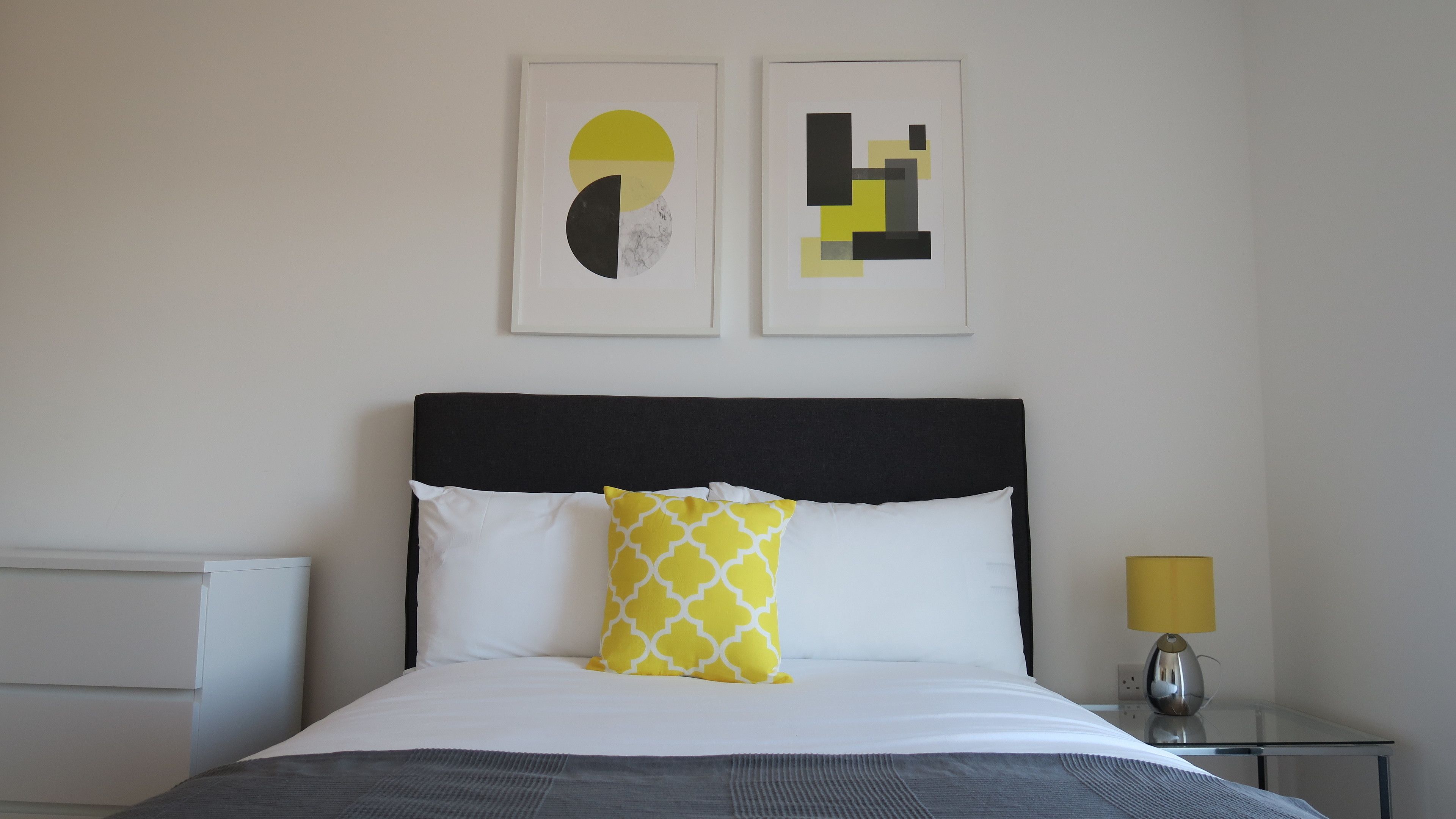 Un lit bien fait, avec une tête de lit noire, des oreillers blancs et jaunes et une couverture grise. Deux œuvres d’art encadrées sont accrochées au mur au-dessus du lit.