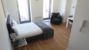 A modern hotel room with a large bed, a chair, a desk, and a view of the city through large windows. - Quay Apartments (Salford)