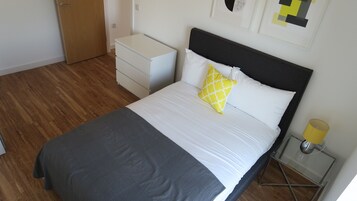 Un dormitorio ordenado con cama, una mesita de noche con lámpara, cómoda y puerta.
