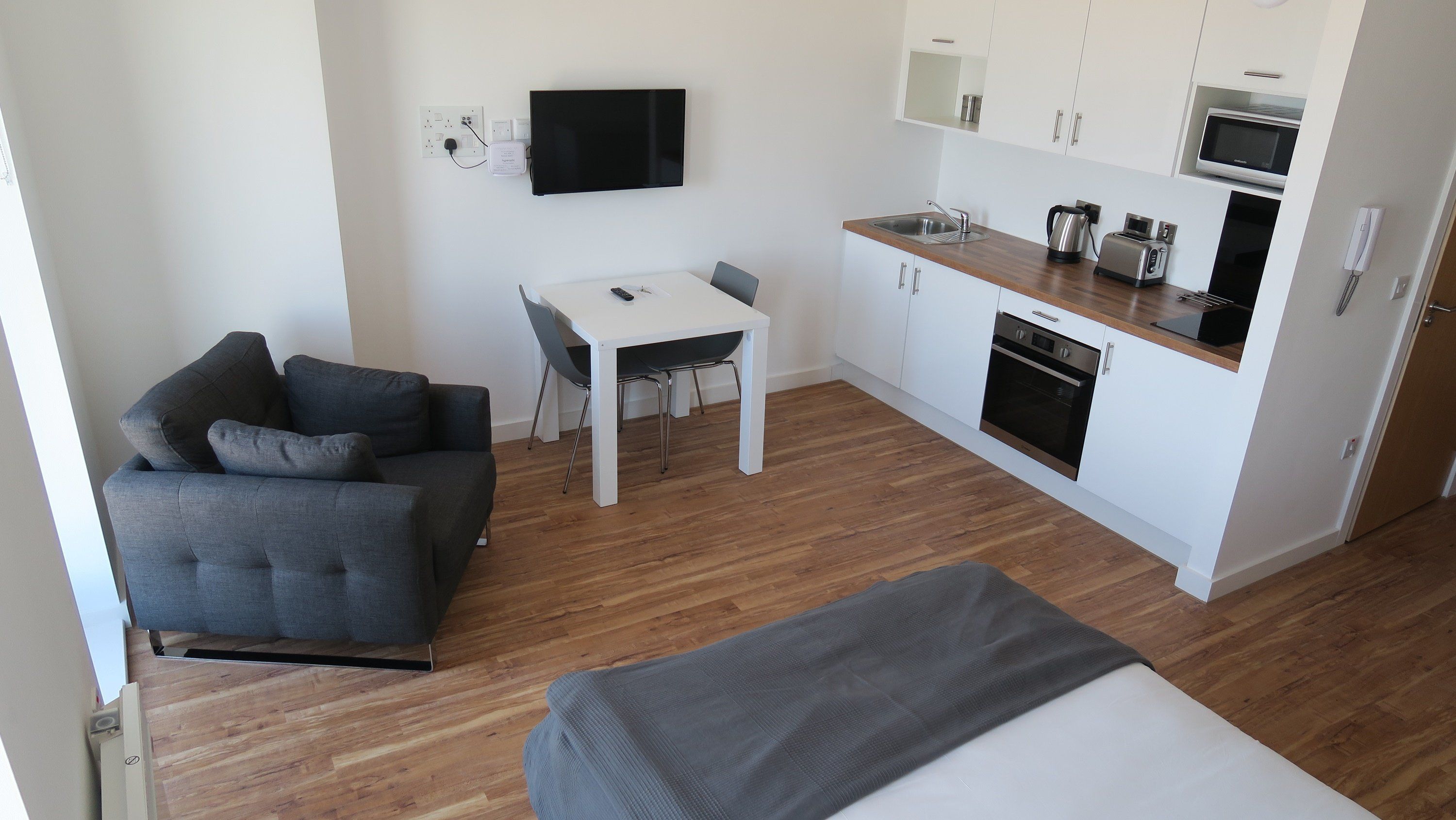 A compact living space with a sofa, a small dining table, a TV mounted on the wall, a kitchenette with a microwave, and an oven.