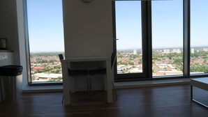 A room with a large window offering a city view, a white desk, and a stainless steel object on the floor. - Quay Apartments (Salford)
